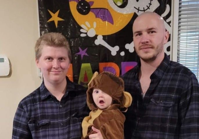 Megan, Colby and their 12-month-old daughter, wearing a monkey costume, pose in front of a Halloween banner.